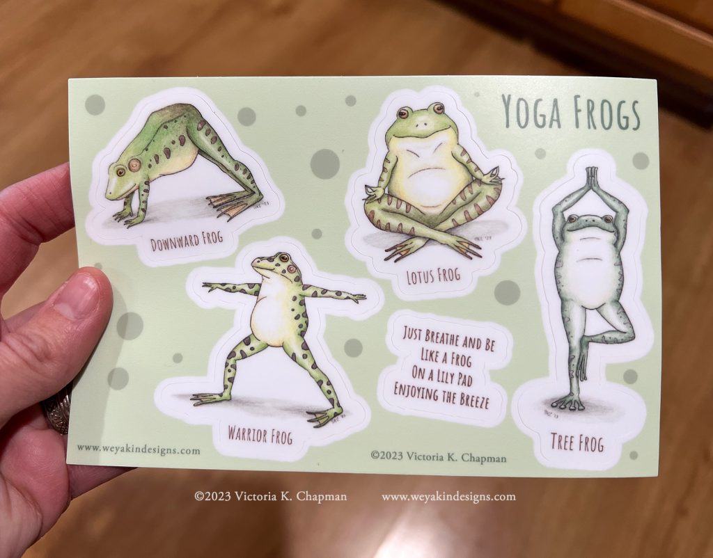 A hand holds a sticker sheet of frogs doing yoga. Art by Victoria K. Chapman.