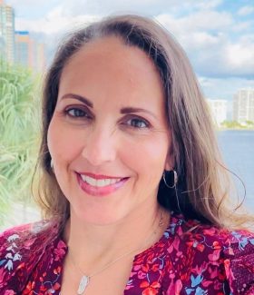 A headshot of Angela Quezada Padron. She has long brown hair, brown eyes and is smiling while wearing a pink and purple floral blouse. There are palm trees and a city in the background.