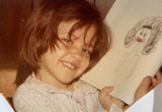 A photo of Angela Quezada Padron as a little girl, smiling and holding up a picture she drew.