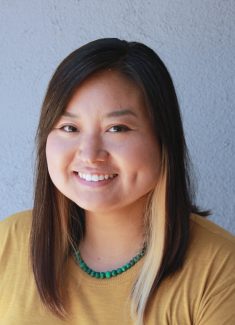 A photo of children's illustrator, Shiho Pate, smiling for the camera. She wears a mustard colored shirt, turquoise necklace, and has shoulder-length dark hair with highlights.