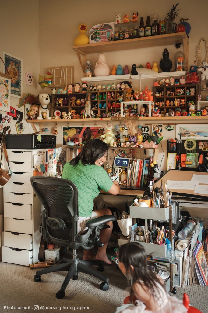 Shiho Pate sitting in her studio at her desk with her daughter playing behind her. The room is filled with toys and fun art.